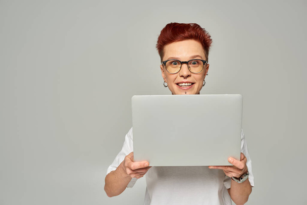 erstaunte queere Freelancerin in weißem T-Shirt und Brille, Laptop in der Hand und Kamera auf grau - Foto, Bild