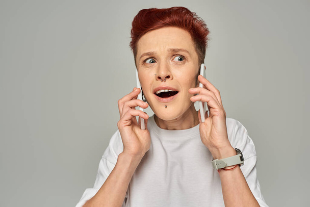 onder de indruk en verbaasd roodharige homo met open mond praten op smartphones op een grijze achtergrond - Foto, afbeelding