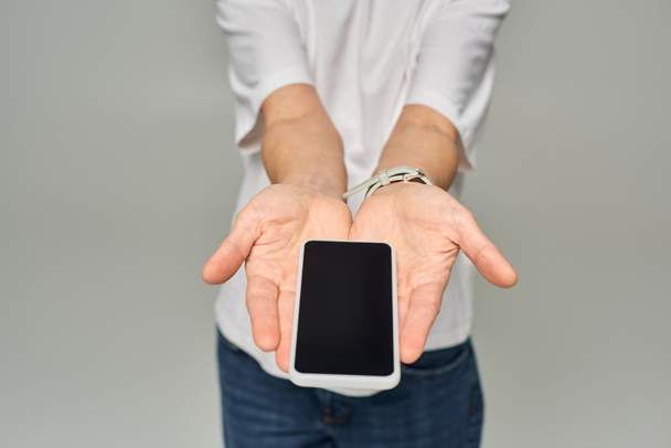 vista recortada de la persona que sostiene el teléfono móvil con pantalla en blanco sobre fondo gris, teléfono inteligente en las manos - Foto, Imagen
