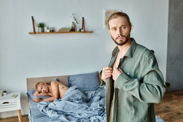 разочарованный бородатый гей одевается рядом с парнем спит в спальне по утрам, любовный конфликт - Фото, изображение