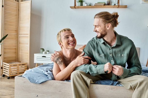 joyful tattooed gay man looking at smiling bearded boyfriend sitting in bedroom, happy relationship - Foto, afbeelding