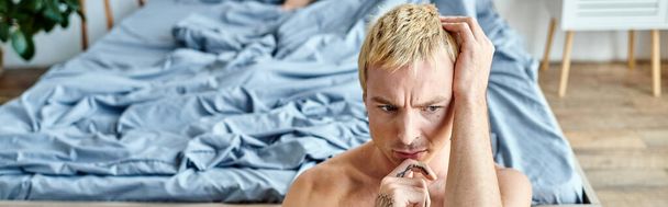 przemyślany i samotny wytatuowany mężczyzna z blond farbowanymi włosami w pobliżu dużego łóżka w sypialni w domu, baner - Zdjęcie, obraz