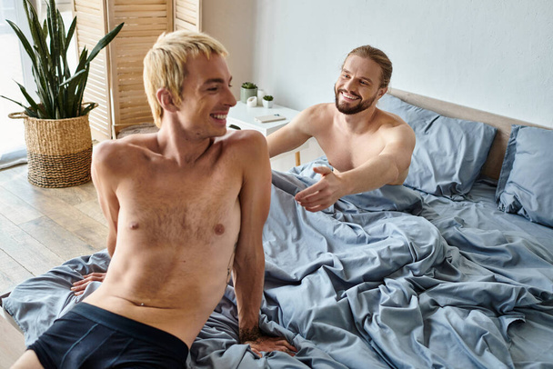 happy bearded gay man with outstretched hands near smiling boyfriend sitting on bed in morning - Foto, afbeelding