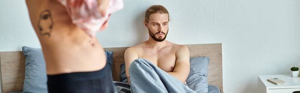 засмучений бородатий гей чоловік сидить на ліжку поруч з любовним партнером, одягненим у спальню, горизонтальний банер - Фото, зображення