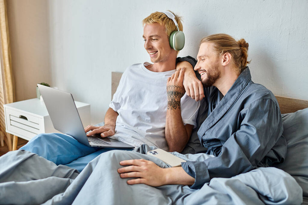 cheerful gay man in headphones using laptop near bearded boyfriend with smartphone in bedroom - Foto, immagini