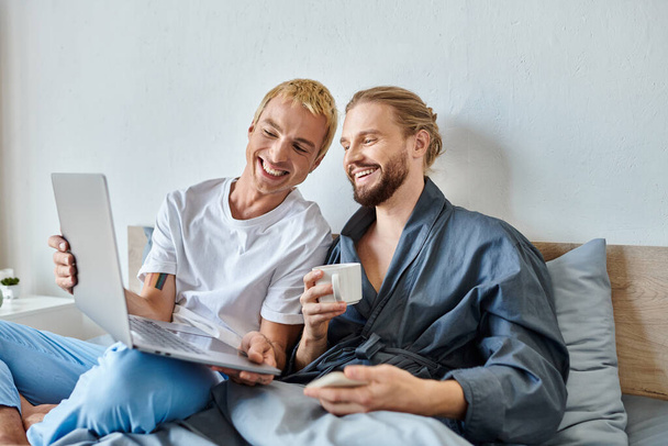 ベッドルームで朝のコーヒーを飲む笑顔のボーイフレンドにノートパソコンを示す陽気なゲイ男性 - 写真・画像