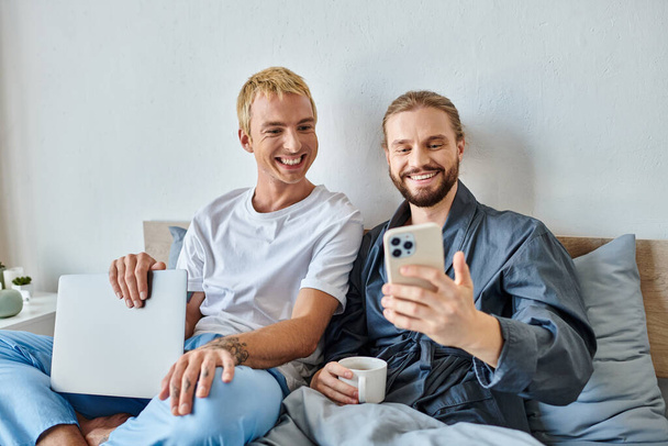 radosny gej człowiek z kawa filiżanka pokazując smartfon dla uśmiechnięty chłopak z laptop w sypialnia - Zdjęcie, obraz