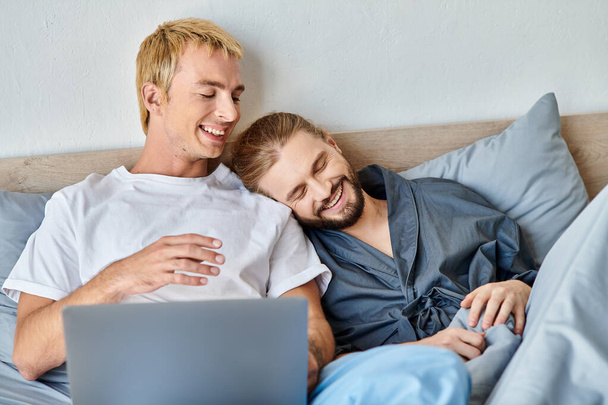улыбающаяся гей-пара смотрит романтический фильм на ноутбуке в спальне, счастливого утреннего отдыха на кровати - Фото, изображение