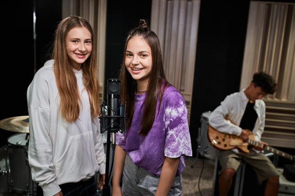 fröhliche hübsche Teenager-Mädchen schauen in die Kamera neben dem Mikrofon, während ihr Freund Gitarre spielt - Foto, Bild