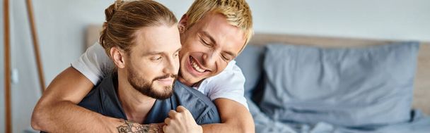 radostný tetovaný gay muž objímající vousatého přítele usmívajícího se v ranní ložnici, horizontální prapor - Fotografie, Obrázek