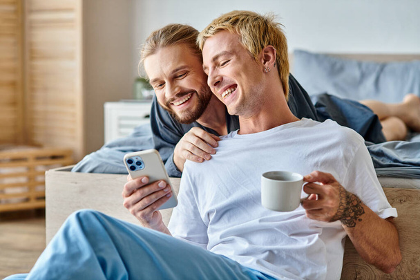 wesoły brodaty gej człowiek uśmiech w pobliżu chłopak z kawa filiżanka za pomocą telefon komórkowy w sypialnia - Zdjęcie, obraz