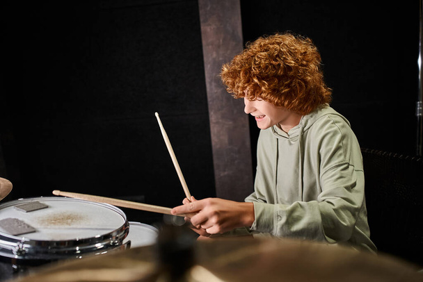 χαρούμενο ταλαντούχο έφηβο αγόρι με κόκκινα μαλλιά σε casual άνετη ενδυμασία παίζοντας τύμπανα στο στούντιο - Φωτογραφία, εικόνα