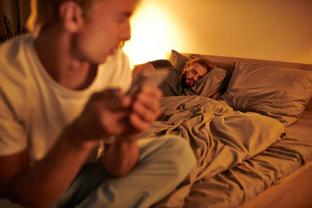 gay muž spí v noci, zatímco nevěrný přítel chatování na smartphone na rozmazané popředí - Fotografie, Obrázek
