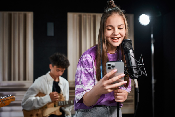 lustiges entzückendes Teenager-Mädchen, das neben ihrem verschwommenen Gitarristen singt und auf das Smartphone schaut - Foto, Bild