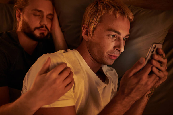 nielojalny gej człowiek przeglądanie data aplikacja na telefon komórkowy w pobliżu chłopak śpi w nocy w sypialnia - Zdjęcie, obraz
