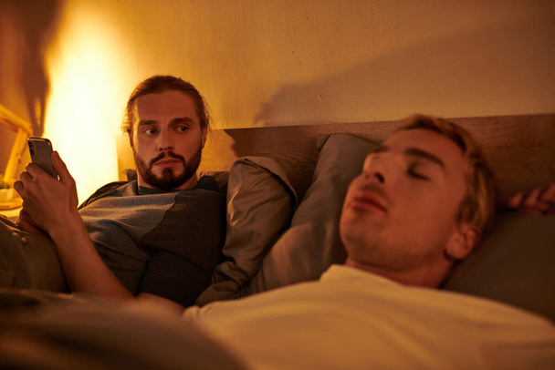 niewierny brodaty gej wiadomości na telefon komórkowy w pobliżu śpiący chłopak w nocy w sypialnia - Zdjęcie, obraz