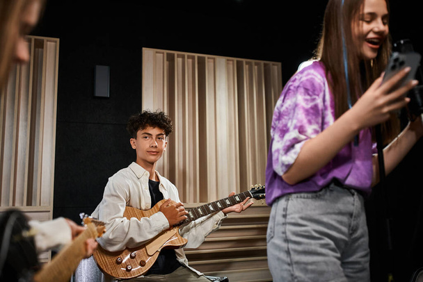 entzückender netter Teenager, der Gitarre spielt und aufmerksam auf seine Freunde schaut, Musikgruppe - Foto, Bild
