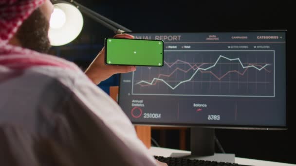 Man met een groene scherm mobiele telefoon terwijl hij naar zakelijke jaarverslagen op de computer kijkt. Zelfstandige met chroma sleutel mobiele telefoon in de hand oplossen van taken thuis - Video