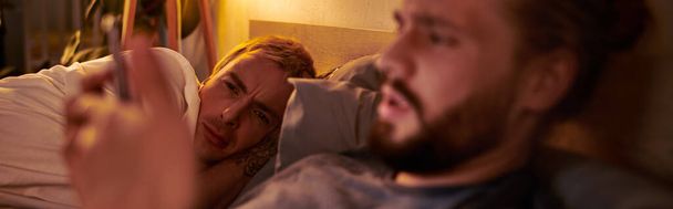 disloyal gay man messaging on smartphone near discouraged boyfriend at night in bedroom, banner - Photo, Image