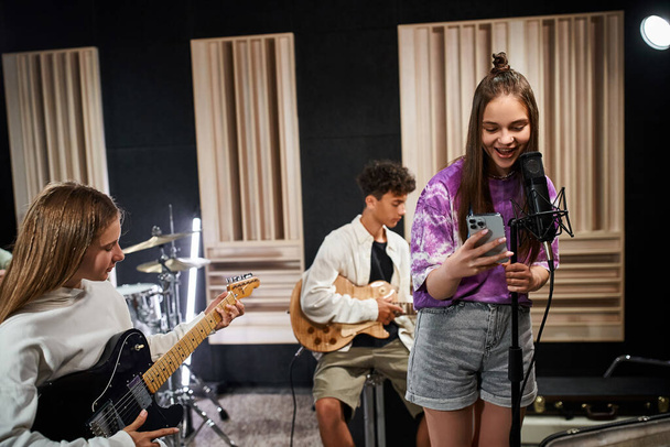 χαρούμενο έφηβο κορίτσι που κοιτάζει το τηλέφωνο και τραγουδά δίπλα στους φίλους της παίζοντας κιθάρες, μουσικό συγκρότημα - Φωτογραφία, εικόνα
