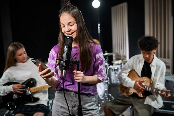 zaměřit se na veselý dospívající dívka v živé oblečení zpěv a při pohledu na telefon v blízkosti její rozmazané kytaristy - Fotografie, Obrázek