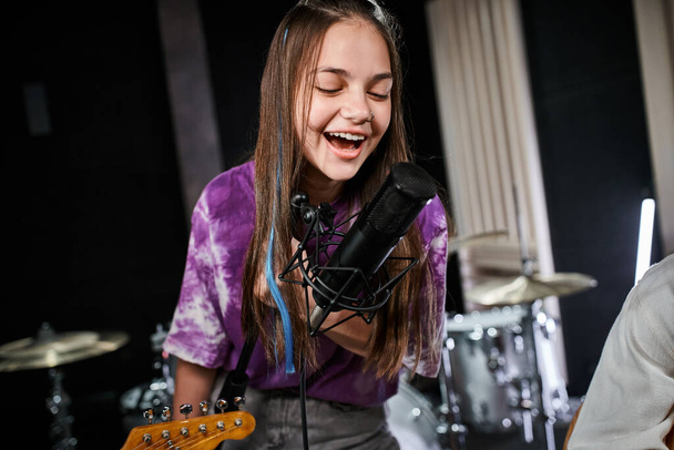 χαρούμενο αξιολάτρευτο έφηβο κορίτσι σε ζωντανή ενδυμασία τραγουδώντας ενεργά στο μικρόφωνο, ενώ στο στούντιο - Φωτογραφία, εικόνα