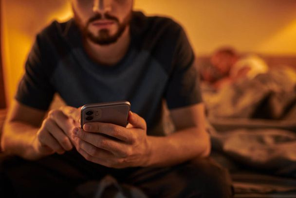 abgeschnittene Ansicht untreuer schwuler Mann auf Smartphone in der Nähe von Partner, der nachts im Schlafzimmer schläft - Foto, Bild