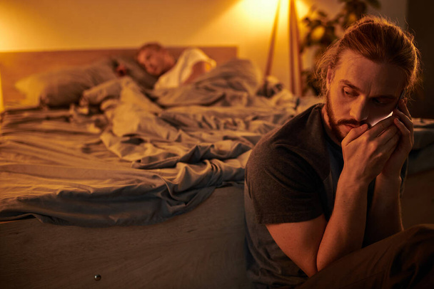 лояльний бородатий гей чоловік розмовляє на мобільному телефоні біля сплячого хлопця вночі в спальні - Фото, зображення