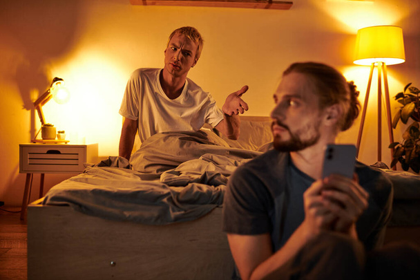 displeased gay man quarreling with boyfriend messaging on smartphone at night in bedroom, trouble - Foto, afbeelding