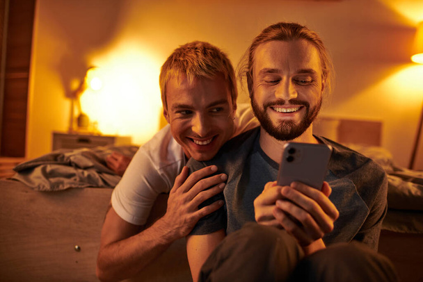 夜の寝室で笑顔のボーイフレンドの近くで携帯電話でインターネットを閲覧する楽しいゲイの男 - 写真・画像