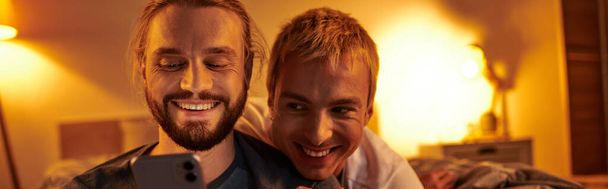 夜の寝室で笑顔のボーイフレンドの近くの携帯電話でインターネットを閲覧する楽しいゲイ男性, バナー - 写真・画像