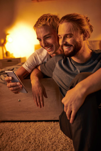 χαρούμενη γενειοφόρος ομοφυλόφιλος άνδρας περιήγηση στο διαδίκτυο στο κινητό τηλέφωνο κοντά χαμογελαστό φίλο στο υπνοδωμάτιο τη νύχτα - Φωτογραφία, εικόνα