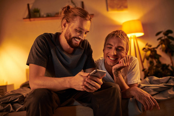 happy bearded gay man browsing internet on mobile phone near smiling boyfriend in bedroom at night - Photo, Image
