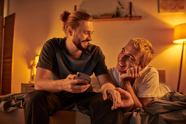 Счастливый бородатый гей просматривает интернет по мобильному телефону рядом с улыбающимся парнем в спальне ночью - Фото, изображение