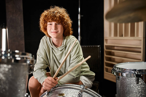 cheerful adorable red haired teenager in casual vivid attire in front of drum set looking at camera - Photo, Image