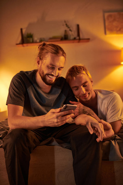 smiling bearded gay man browsing internet on mobile phone near happy boyfriend in bedroom at night - Photo, Image