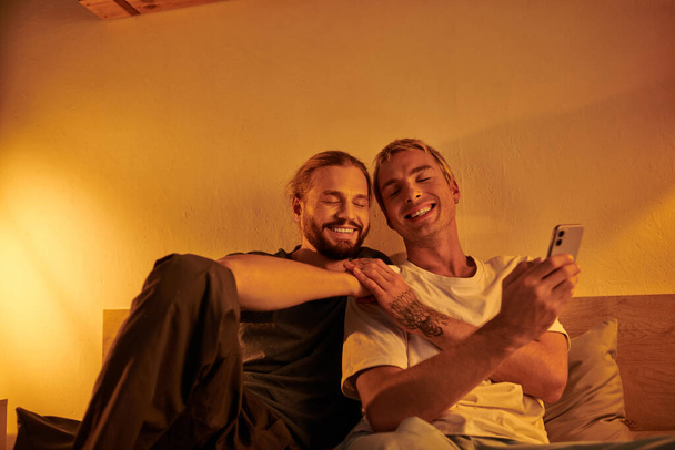 幸せな入れ墨のゲイ男性は夜の寝室で笑顔のひげ付きボーイフレンドの近くにスマートフォンを保持しています - 写真・画像