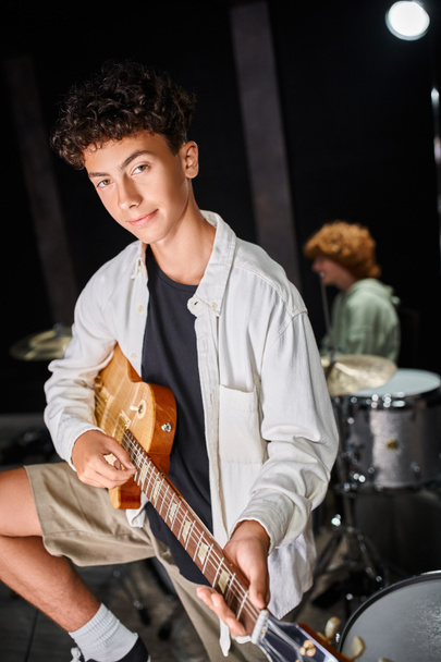 Konzentrierter Teenager in lässiger Kleidung, der in der Nähe seines verschwommenen rothaarigen Schlagzeugers Gitarre spielt - Foto, Bild