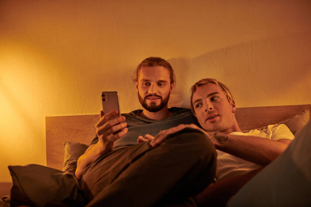 feliz barbudo gay homem navegação social mídia no smartphone perto sorrindo namorado no quarto no noite - Foto, Imagem