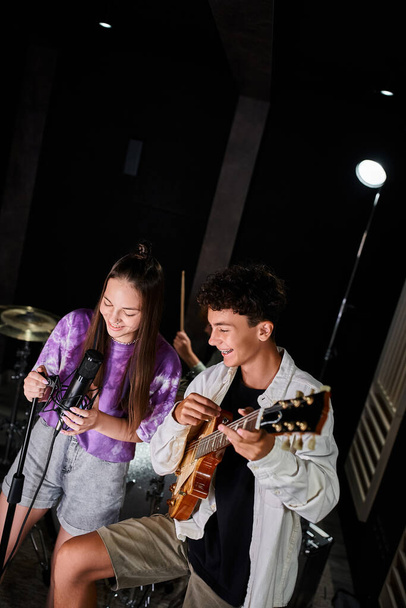 gelukkig getalenteerd tiener meisje in levendige kledij zingen in microfoon naast haar gitarist in de studio - Foto, afbeelding