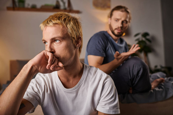 offended gay man holding hand near face and looking away near boyfriend at night in bedroom - Photo, Image