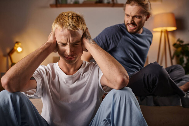 強調されたゲイ男性は夜の寝室で喧嘩している怒っているボーイフレンドの近くに手で耳を覆う - 写真・画像