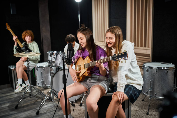 バックドロップにドラムやギターとぼやけた友人とギターを歌う10代の女の子に焦点を当てます - 写真・画像