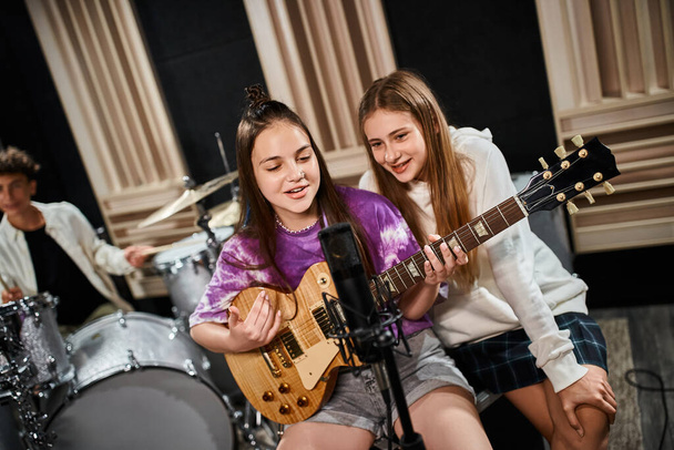 focus on cheerful talented girls playing guitar and singing with their blurred drummer on backdrop - Photo, Image