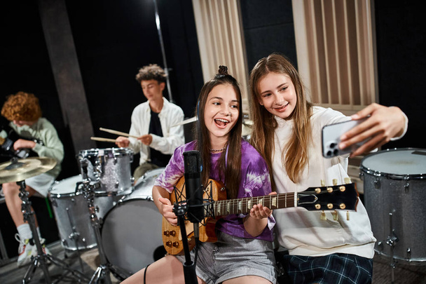 Fokus auf fröhliche Teenager-Mädchen, die Selfies machen und mit verschwommenen Freunden singen, die Instrumente spielen - Foto, Bild