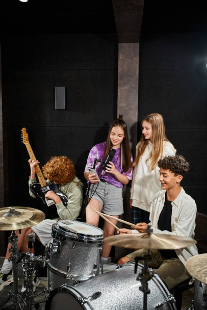 αξιολάτρευτα έφηβα αγόρια που παίζουν όργανα ενώ τα κορίτσια τραγουδούν και κοιτάζουν το τηλέφωνο, μουσική ομάδα - Φωτογραφία, εικόνα