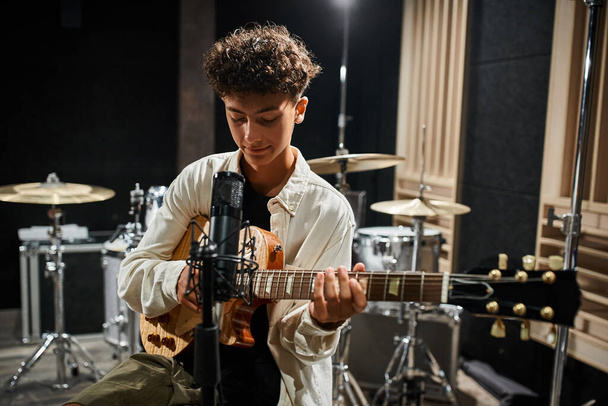 jolly talented teenage boy in comfy casual attire playing his guitar in studio and smiling slightly - Photo, Image