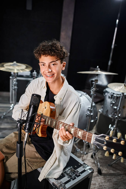 όμορφος έφηβος μουσικός με τιράντες σε casual ενδυμασία παίζοντας κιθάρα και χαμογελώντας στην κάμερα - Φωτογραφία, εικόνα