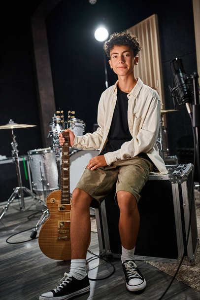 カジュアルな衣装でハンサムな才能のある十代の少年が座ってギターを保持し,カメラを見ている - 写真・画像
