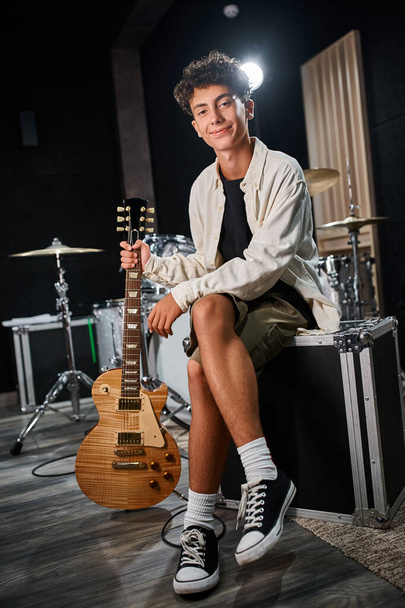 alegre bom olhar adolescente em traje casual segurando guitarra e sorrindo para a câmera em estúdio - Foto, Imagem